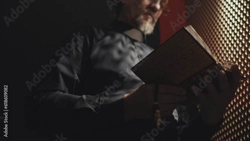 Low angle selective focus shot of mature Catholic priest holding rosary standing in confessional booth opening Bible book photo