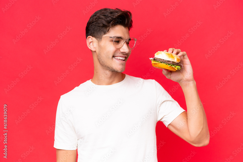 Young Caucasian man holding a burger isolated on red background with happy expression Young Caucasian man holding a burger isolated on red background with happy expression