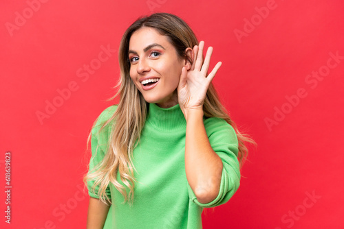 Young Uruguayan woman isolated on red background listening to something by putting hand on the ear