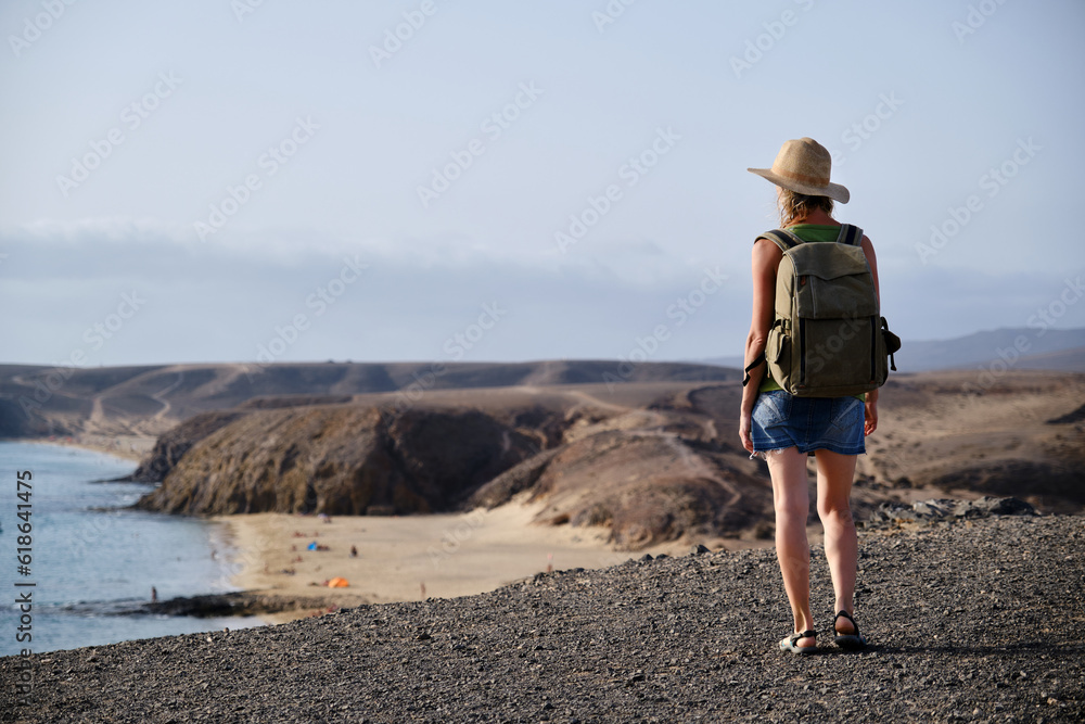 A Woman with hat and backpack, standing on top of a cliff and looking out to sea and beach at sunset. Travel and tourism.