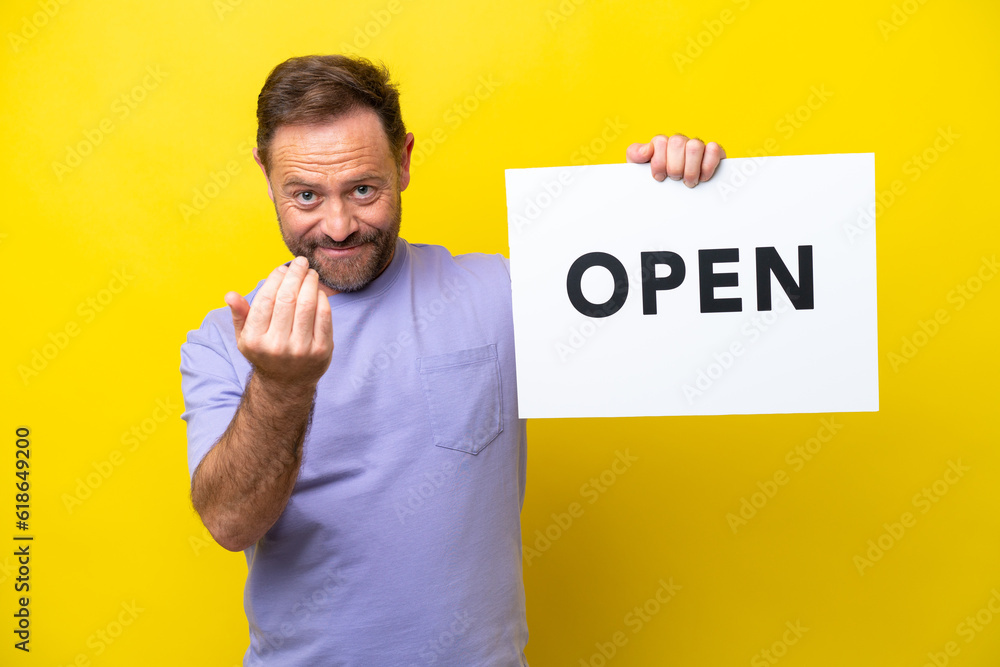 Middle age caucasian man isolated on yellow background holding a placard with text OPEN and doing coming gesture Middle age caucasian man isolated on yellow background holding a placard with text OPEN and doing coming gesture
