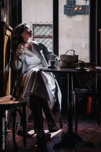 Young girl in white dress drinking coffee in a coffee shop near the window in the sun. Silhouette.