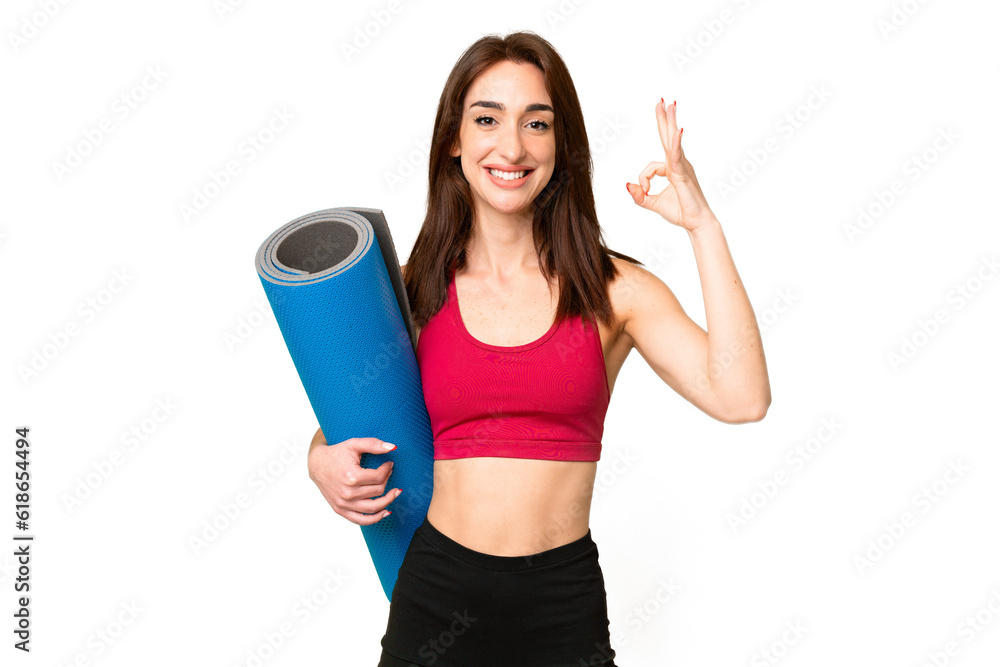 Young sport caucasian woman going to yoga classes while holding a mat over isolated chroma key background showing ok sign with fingers