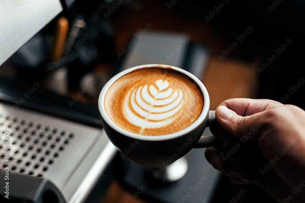 Everyday Coffee Bliss: Hand Holding a Cup of Latte Art with Coffee Maker Machine in the Background, Illuminated by Low Key Light Everyday Coffee Bliss: Hand Holding a Cup of Latte Art with Coffee Maker Machine in the Background, Illuminated by Low Key Light