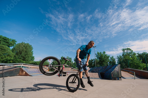 A middle-aged tattooed professional bmx rider is performing freestyle tricks in a skate park.