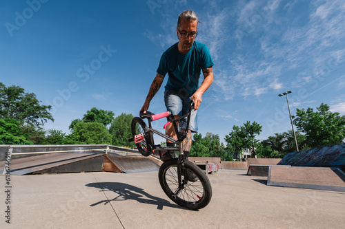 A mature cool tattooed man is practicing tricks and stunts on a bmx in a skate park.