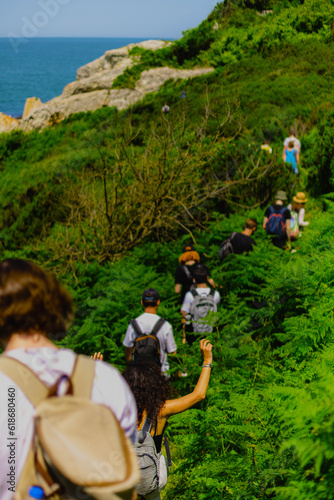 Group of hikers with backpacks wade through thickets of ferns along seashore on sunny summer day