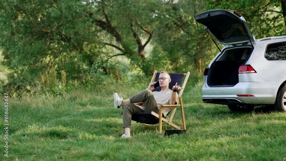 Relaxed male speaks on the phone sitting in the folding chair. Remote work conversation in the woods.