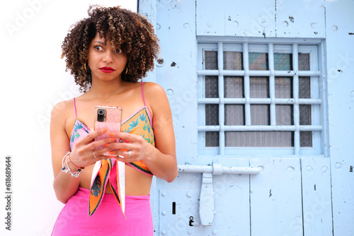Photography A girl with afro hair and dressed in bright colors doesn't seem to like what she sees on her cell phone next to a pretty blue door