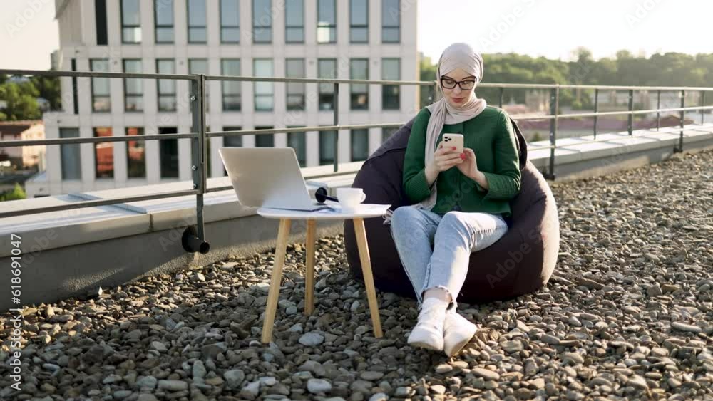 Full length view of charming woman in headscarf holding phone while sitting in beanbag near coffee table with gadgets. Delighted muslim person adoring city view in sunlight on panoramic terrace. Full length view of charming woman in headscarf holding phone while sitting in beanbag near coffee table with gadgets. Delighted muslim person adoring city view in sunlight on panoramic terrace.