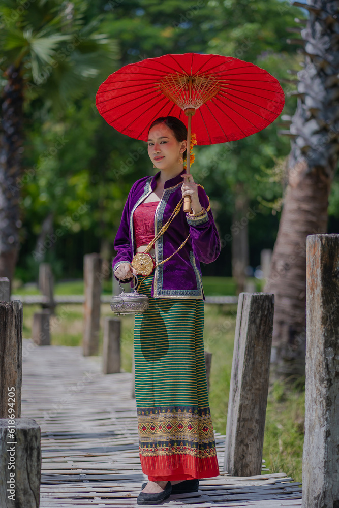 Obraz premium Beautiful pretty young Asian woman wearing a millionaire traditional Thai dress Lanna style standing with an antique silver bag and umbrella in a green natural park.