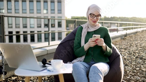 Full length view of charming woman in headscarf holding phone while sitting in beanbag near coffee table with gadgets. Delighted muslim person adoring city view in sunlight on panoramic terrace.