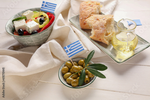 Dishes with ciabatta bread, salad, green olives and flags of Greece on white wooden table