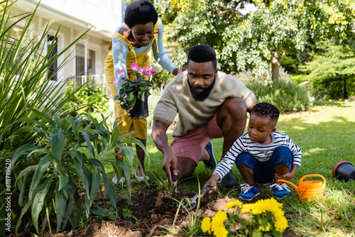 African american woman holding flowers while father and son digging dirt on grassy land in yard