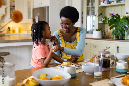 Smiling african american mother cleaning daughter's face with tissue paper at dining table