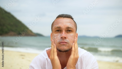 Close-up portrait man apply sun cream protection lotion, looking at camera. Funny man on beach near sea smearing sunscreen cream. Concept facial skin care, travel on tropical island weekend beach sea