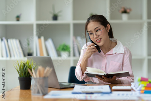 Confident Asian businesswoman using mobile phone to contact customers to talk Negotiate on business matters Happily logging deals while sitting at his desk in the office.