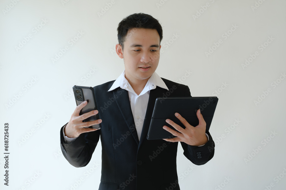 Young Asian businessman holding a smartphone and a tablet device and looking at it. Isolated white background. Young Asian businessman holding a smartphone and a tablet device and looking at it. Isolated white background.