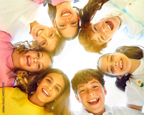 Happy children play together in free time, hug, huddle and have fun. Low angle bottom view from below group best kid friends look down at camera with smiling expressions in dreamy yellow sunlight haze