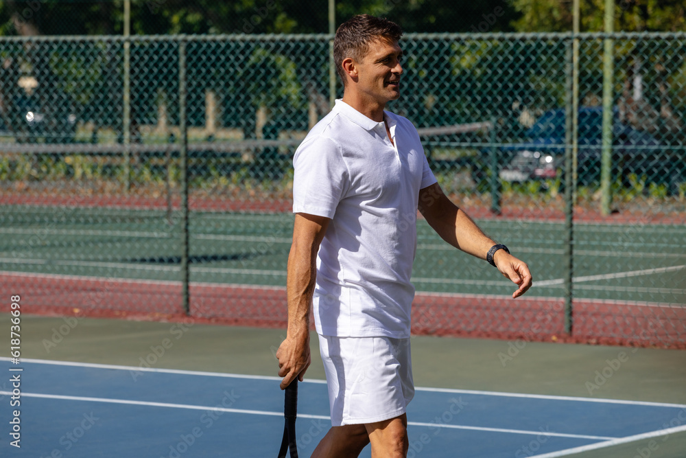 Happy caucasian male tennis player with tennis racket walking on outdoor tennis court on sunny day Happy caucasian male tennis player with tennis racket walking on outdoor tennis court on sunny day