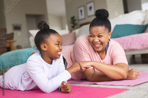 Happy african american mother and daughter lying on yoga mats and fist-bumping in living room photo