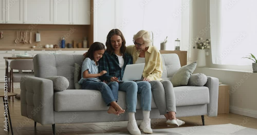 Three attractive multi generational women spend time with wireless laptop relaxing on coach in living room, enjoy internet fun content, laughing on on-line videos, watch movie on streaming services