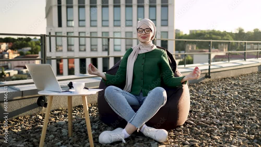 Portrait of muslim woman in headscarf and spectacles taking seated position in pouf chair with fingers in gyan mudra. Joyous lady in cozy outfit improving concentration during remote work outdoors. Portrait of muslim woman in headscarf and spectacles taking seated position in pouf chair with fingers in gyan mudra. Joyous lady in cozy outfit improving concentration during remote work outdoors.
