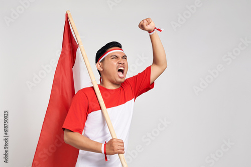 Excited young Asian man standing holding Indonesian flag with raised hands while Celebrate Indonesia independence day on 17 August isolated over white background