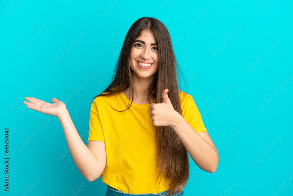 Young caucasian woman isolated on blue background holding copyspace imaginary on the palm to insert an ad and with thumbs up Young caucasian woman isolated on blue background holding copyspace imaginary on the palm to insert an ad and with thumbs up