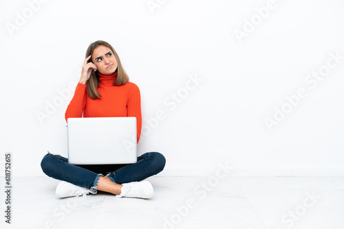 Young caucasian woman sitting on the floor with a laptop having doubts and with confuse face expression