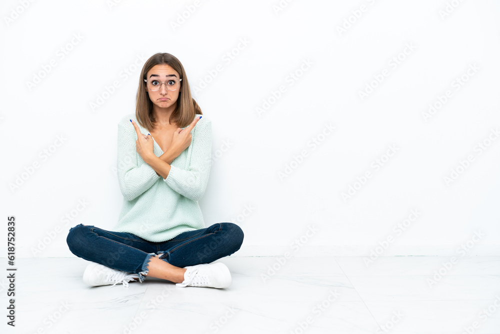 Young caucasian woman sitting on the floor isolated on white background pointing to the laterals having doubts Young caucasian woman sitting on the floor isolated on white background pointing to the laterals having doubts