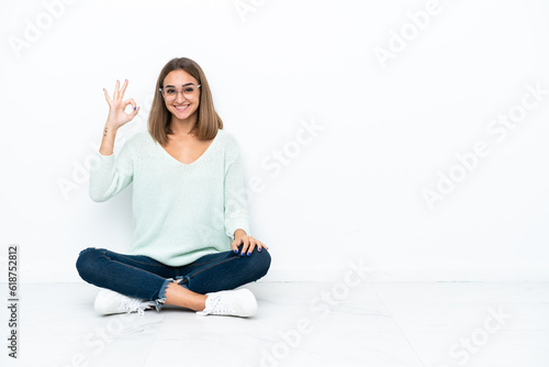 Young caucasian woman sitting on the floor isolated on white background showing ok sign with fingers