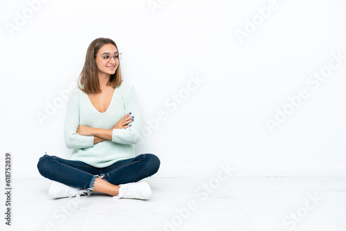 Young caucasian woman sitting on the floor isolated on white background with arms crossed and happy