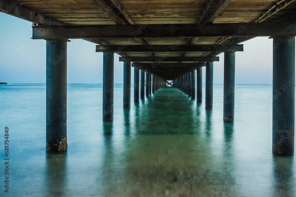 Metal supports of the pier at dawn. View of the pillars under the wooden pier in the sea. Long exposure of the sea as a haze