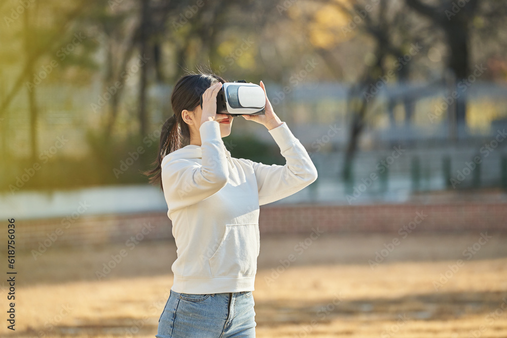 Young college student female model wearing 3D glasses and experiencing virtual reality in an outdoor autumn lawn plaza at a university in South Korea, Asia.