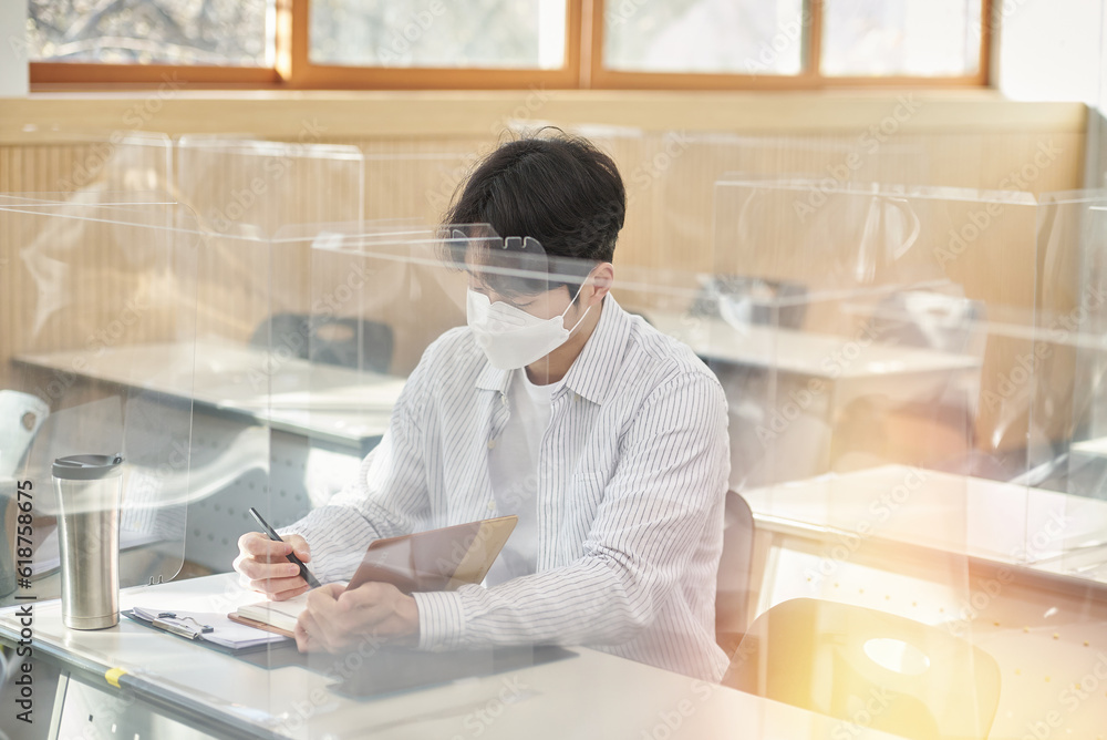 A young male Asian college student is learning or listening to a lesson while her face is outside a protective shield on the desk in a university classroom in Korea during the pandemic. A young male Asian college student is learning or listening to a lesson while her face is outside a protective shield on the desk in a university classroom in Korea during the pandemic.