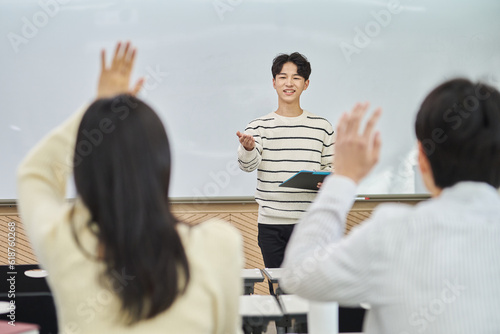 An Asian young man is standing in front of a lecture hall at a university in South Korea, giving a presentation or lecture. In front of him are male and female students. 