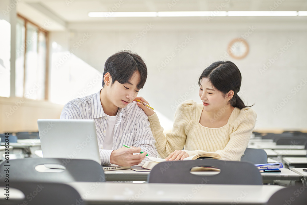 Fototapeta premium In a higher education classroom in South Korea, young university students are listening to a lecture, studying, and talking. A woman and a man are in the background