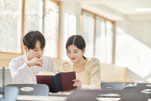 In a higher education classroom in South Korea, young university students are listening to a lecture, studying, and talking. A woman and a man are in the background