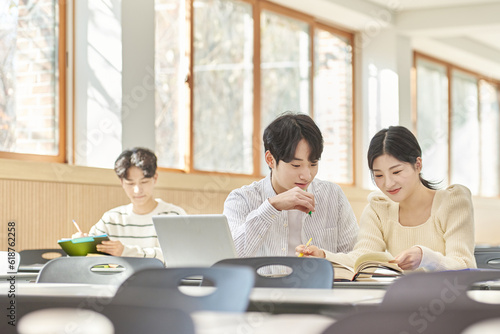 In a higher education classroom in South Korea, young university students are listening to a lecture, studying, and talking. A woman and a man are in the background