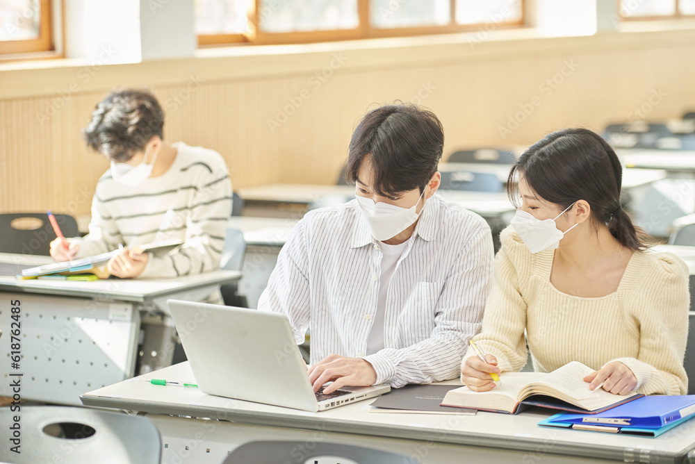 In a higher education classroom in South Korea, young university students wearing masks are listening to a lecture, studying, and talking. A woman and a man are in the background In a higher education classroom in South Korea, young university students wearing masks are listening to a lecture, studying, and talking. A woman and a man are in the background