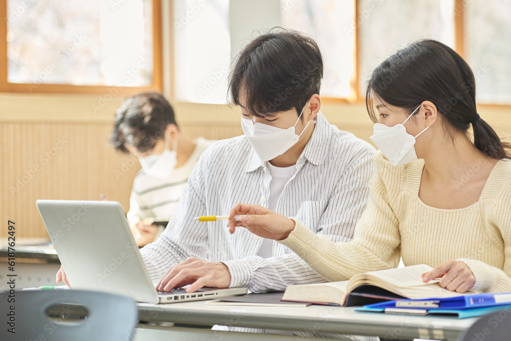 In a higher education classroom in South Korea, young university students wearing masks are listening to a lecture, studying, and talking. A woman and a man are in the background