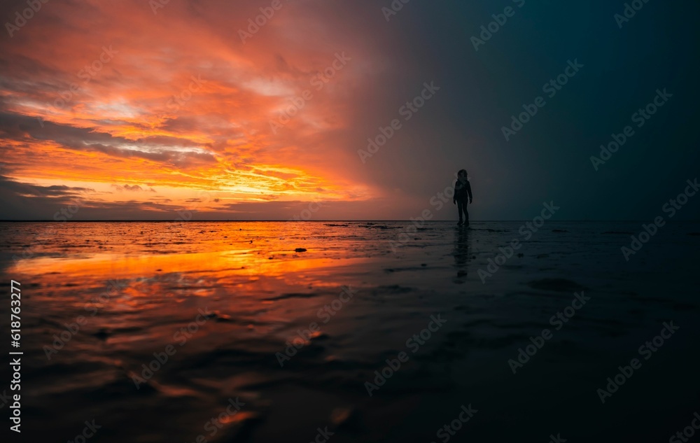 Silhouette of a person standing in shallow water against the sea at orange sunset