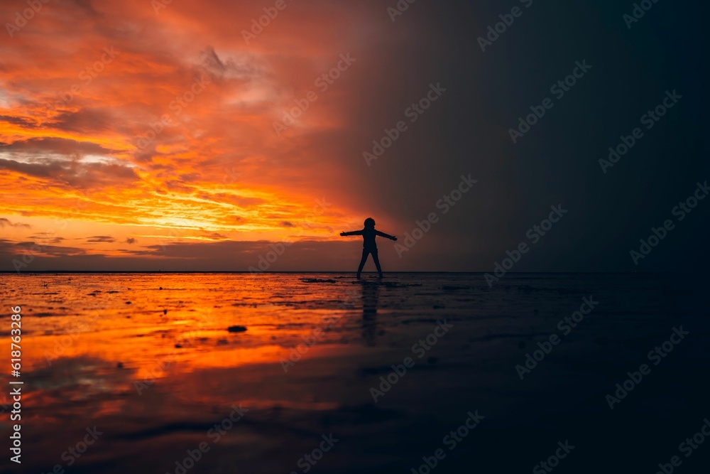 Silhouette of a person standing in shallow water against the sea at orange sunset