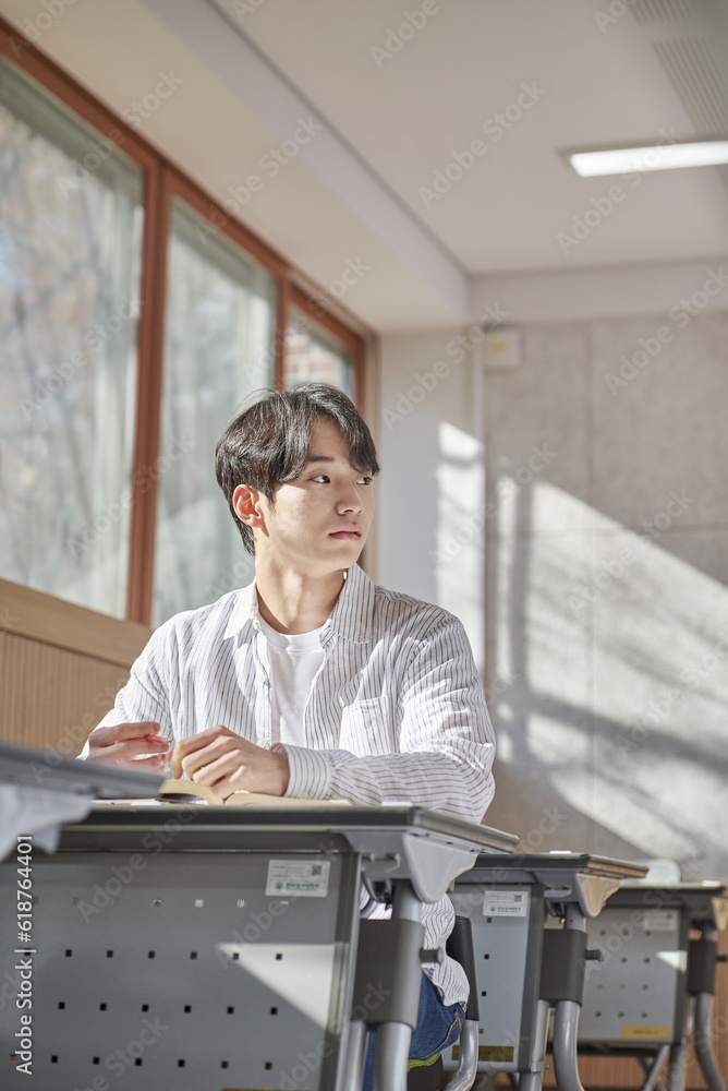 Fototapeta premium A young college student is sitting at a desk in a university classroom in South Korea, Asia. He is either reading a book or looking out of the window