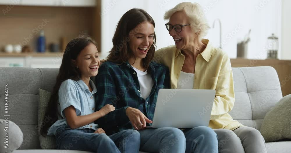 Mature granny spend time with preteen grandchild and millennial daughter laugh while watch funny on-line content, movie via streaming services sit on sofa with laptop. Amusement, family leisure, tech Mature granny spend time with preteen grandchild and millennial daughter laugh while watch funny on-line content, movie via streaming services sit on sofa with laptop. Amusement, family leisure, tech