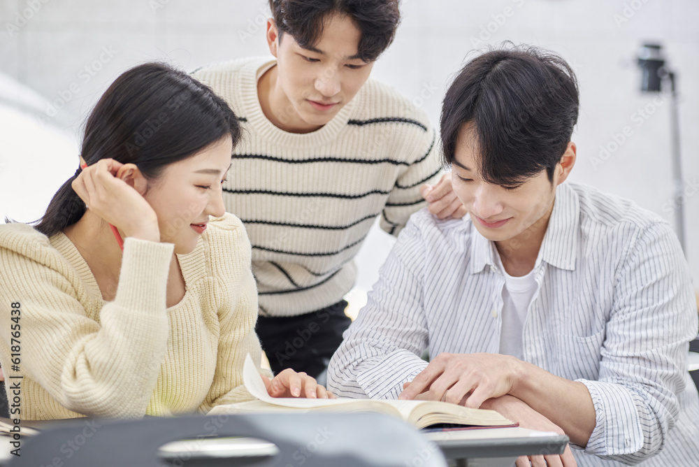 Three young male and female college students models sitting or standing at desks in a university classroom in South Korea, Asia, talking or having a discussion Three young male and female college students models sitting or standing at desks in a university classroom in South Korea, Asia, talking or having a discussion