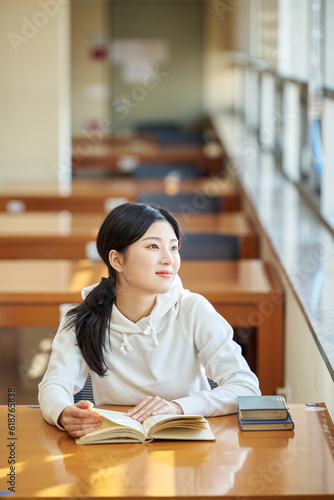 Asian Korean University Library Young female college student model sitting at a desk by a window with light coming in, reading a book, looking out the window