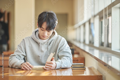 Asian Korean University Library Young male college student model sitting at a desk by a window with light coming in, reading a book, looking out the window or leaning down.