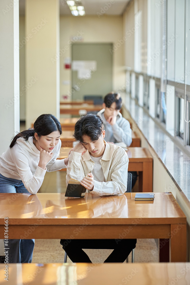 Three college students, both male and female, are sitting or standing at a desk in a library at a South Korean university in Asia, looking at laptops and books while having a conversation. Three college students, both male and female, are sitting or standing at a desk in a library at a South Korean university in Asia, looking at laptops and books while having a conversation.